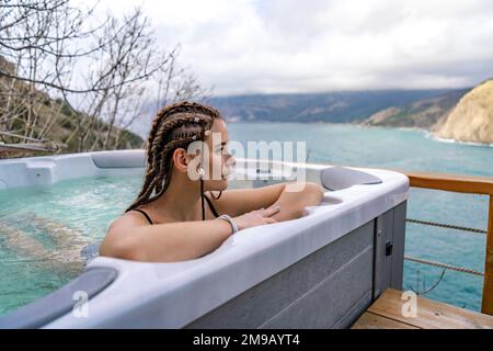 Nehmen Sie sich Zeit für sich. Badesachen im Freien mit Blick auf die Berge und das Meer. Eine Frau im schwarzen Badeanzug entspannt sich im Hotelpool und bewundert die Aussicht Stockfoto