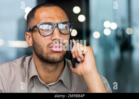 Ein seriöser und fokussierter afroamerikanischer Geschäftsmann, der mitten im Büro telefoniert, hört einem Gesprächspartner in einem lässigen Hemd und einer Brille zu. Stockfoto