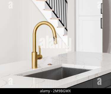 Ein Küchenwaschbecken Detail aufgenommen mit einem goldenen Wasserhahn, graue Insel, weißen Marmor Arbeitsplatte, und Lichter hängen über der Insel. Stockfoto