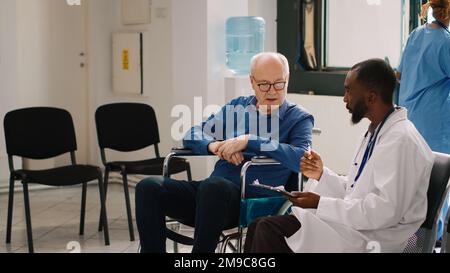 Hausarzt, der sich im Rollstuhl mit älteren Menschen beraten lässt, medizinische Berichtspapiere in der Krankenhauslobby ausfüllt. Patient mit chronischer Behinderung im Wartezimmer im Gespräch mit dem Arzt. Stockfoto