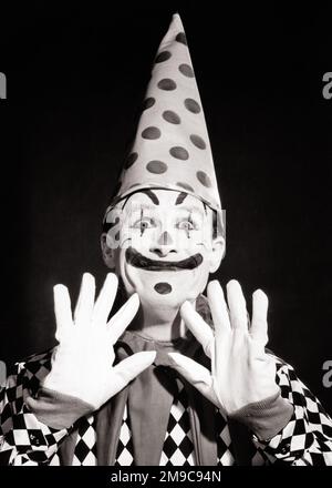 1950S 1960S LÄCHELNDER CLOWN MIT LUSTIGEM GESICHT, DER POLKA DOT DUNCE CAP TRÄGT, ERHOBENE HÄNDE IN WEISSEN HANDSCHUHEN - C3085 DEB001 HARS ERFREULICHE LEBENSSTIL-JOBS STUDIO-AUFNAHME KOPIE-PERSONEN CHARAKTER MÄNNLICHE SCHWARZWEISS-DUMMKOPF BREITE CLOWNS AUGENKONTAKT BIZARR DOT GESCHICKLICHKEIT WANZAUGEN HUMOR BERUF GLÜCK FÄHIGKEITEN SELTSAMER KOPF UND SCHULTERN FRÖHLICHE DARSTELLER POLKA GROTESKE AUFREGUNG BILDEN KOMISCHE UNKONVENTIONELLE ENTERTAINER BERUFE LÄCHELN COULROPHOBIA KONZEPTIONELLE KOMÖDIE JESTER FREUDVOLL VERRÜCKTER DEB001 IDIOSYNKRATISCH MIT BREITEN AUGEN AMÜSANTE KREATIVITÄT EXZENTRISCH ERSCHRECKT JUNGER ERWACHSENER SCHWARZ UND WEISS Stockfoto