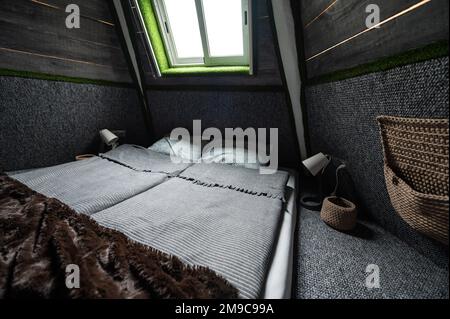 Doppelbett auf Teppichboden im Schlafzimmer der Holzkabine. Pyramidenförmiger Bungalow von innen. Kleines Zimmer, abgewinkelte Wände, Fensterbeleuchtung, Bett gemacht. Handgefertigt Stockfoto