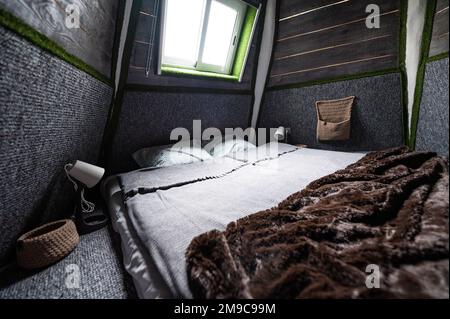 Doppelbett auf Teppichboden im Schlafzimmer der Holzkabine. Pyramidenförmiger Bungalow von innen. Kleines Zimmer, abgewinkelte Wände, Fensterbeleuchtung, Bett gemacht. Handgefertigt Stockfoto