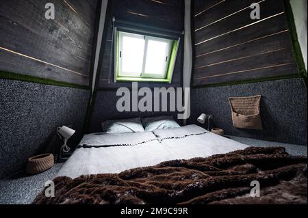 Doppelbett auf Teppichboden im Schlafzimmer der Holzkabine. Pyramidenförmiger Bungalow von innen. Kleines Zimmer, abgewinkelte Wände, Fensterbeleuchtung, Bett gemacht. Handgefertigt Stockfoto