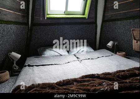 Doppelbett auf Teppichboden im Schlafzimmer der Holzkabine. Pyramidenförmiger Bungalow von innen. Kleines Zimmer, abgewinkelte Wände, Fensterbeleuchtung, Bett gemacht. Handgefertigt Stockfoto