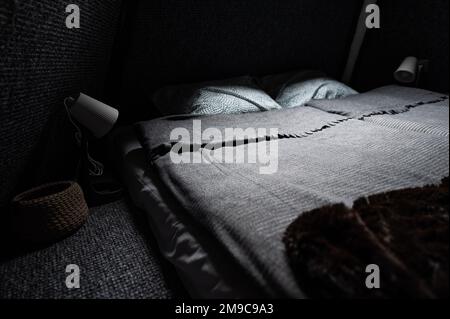 Doppelbett auf Teppichboden im Schlafzimmer der Holzkabine. Pyramidenförmiger Bungalow. Kleines Zimmer, abgewinkelte Wände, Fensterbeleuchtung, Bett, Nachtlampe und Knitte Stockfoto