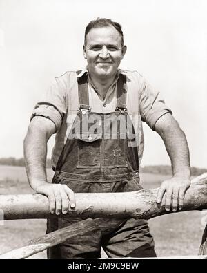 1940S-PORTRÄT EINES STARKEN, SELBSTBESESSENEN BAUERN, DER EINEN LATZANZUG AUS DENIM TRÄGT UND AUF DIE KAMERA SCHAUT, SICH AUF EINEN HOLZZAUN LEHNT UND LÄCHELT - F793 HAR001 HARS GESUNDHEIT KOPIE RAUM HALBE LÄNGE KÖRPERLICHE FITNESS PERSONEN OVERALLS FÜRSORGLICHE LANDWIRTSCHAFT MÄNNER VERTRAUEN DENIM AUSDRÜCKE LANDWIRTSCHAFT SCHWARZWEISS-AUGE KONTAKTFREIHEIT ERFOLG SCHIENE GLÜCK WELLNESS FRÖHLICHE STÄRKE EHRLICHER MUT BAUERN WISSEN FÜHRUNG MÄCHTIGER STOLZ AUF AUTORITÄT BERUFE LÄCHELN KONZEPTIONELL FRÖHLICH STYLISCHE BIB OVERALLS BIB-WACHSTUM MITTE-ERWACHSENER MANN ZUSAMMENGEHÖRIGKEIT SCHWARZ-WEISS WEISSE ETHNISCHE ZUGEHÖRIGKEIT HAR001 ALTMODISCH Stockfoto