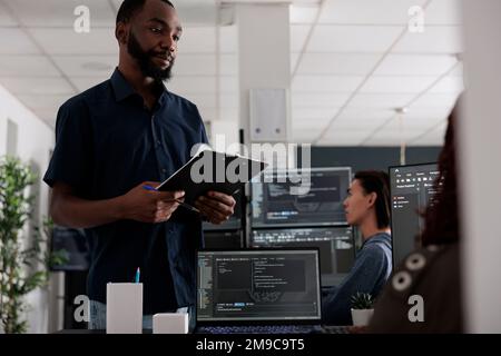 Softwareentwickler, der die Zwischenablage hält, prüft HTML-Code im Terminalfenster, wobei die Serverdatenbank auf dem Computer verwendet wird. Programmierer sprechen über Programmiersprache und Cloud-Computing-Benutzeroberfläche. Stockfoto