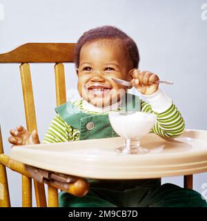 1960S LÄCHELNDES AFROAMERIKANISCHES BABY, DAS EIS ISST, SITZT IM HOCHSTUHL UND TRÄGT EIN GRÜN GESTREIFTES OUTFIT - KB10134 PHT001 HARS-KLEINKIND HAPPY JOY LIFESTYLE ZUFRIEDENHEIT GESTREIFT STUDIO SHOT GESUNDHEIT HOME LIFE COPY SPACE MÄNNER OUTFIT HAPPY HOCHSTUHL KOPF UND SCHULTERN FRÖHLICH AFROAMERIKANER AFROAMERIKANER AUFREGUNG ERNÄHRUNG SCHWARZE ETHNISCHE ZUGEHÖRIGKEIT LÄCHELN KONSUM FREUDIGE ERNÄHRUNG BABY JUNGE ZUSAMMENARBEIT WACHSTUM JUGENDLICHE ALTMODISCHE AFROAMERIKANER Stockfoto