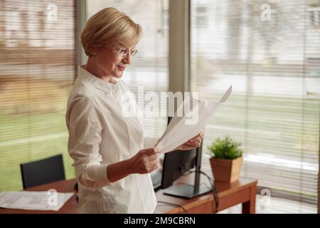 Eine ziemlich reife Freiberuflerin arbeitet mit Dokumenten, während sie im Hintergrund des Home Office steht Stockfoto