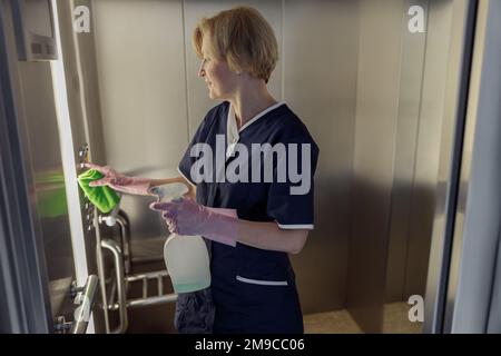 Professionelles Zimmermädchen trägt Uniform und Handschuhe, reinigt den Aufzug mit Reinigungsmittel und Lappen Stockfoto