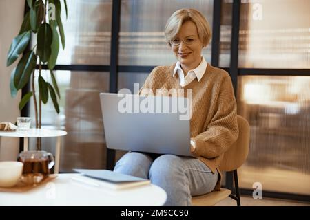 Lächelnde, reife Geschäftsfrau mittleren Alters, die in gemütlicher Zusammenarbeit mit Laptops arbeitet und im Internet surft Stockfoto