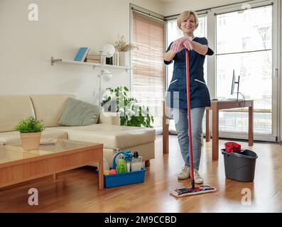 Professionelles lächelndes Hausmädchen im gleichmäßigen Waschboden mit Mopp im Wohnzimmer Stockfoto