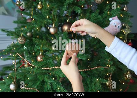 Die Hände des Kindes halten eine Weihnachtsbaumschmuck in Form eines glänzenden Herzens vor dem Hintergrund eines dekorierten Weihnachtsbaums. Stockfoto