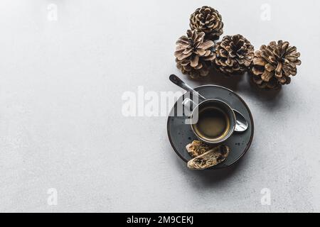 Top-down-Shot einer Tasse Espresso-Kaffee auf einer Untertasse mit kleinem Löffel und zwei Keksen neben dekorativen Kiefernzapfen auf einem Steintisch. Herbstsonnengetränk. Horizontale Aufnahme. Hochwertiges Foto Stockfoto