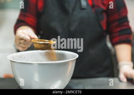 Weibliche Bäckerin mit schwarzer Schürze, die Kakaopulver in eine weiße Schüssel saugt, um Schokoladen-Cupcake-Teig zuzubereiten. Backvorgang. Horizontale Nahaufnahme. Hochwertiges Foto Stockfoto