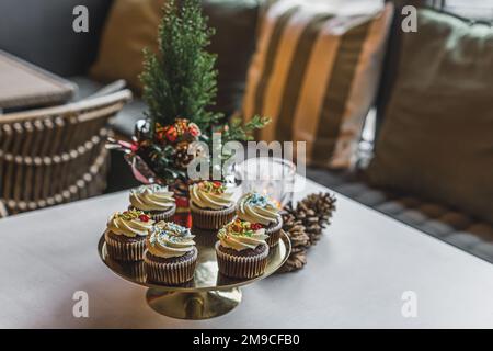 Dekorative festliche Cupcakes, serviert auf einer Goldplatte auf einem Bäckereitisch des Coffeeshops mit kleiner weihnachtsbaumdekoration. Gemütlicher Winterbereich. Winterdesserts. Horizontale Indoor-Aufnahme. Hochwertiges Foto Stockfoto