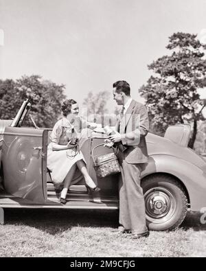 1930S LÄCHELNDE FRAU, DIE DIE KAMERA HÄLT, SITZT IN DER OFFENEN TÜR DES CABRIOS UND SPRICHT MIT EINEM MANN, DER EINEN PICKNICKKORB IN DER HAND HÄLT THERMOS - M4699 HAR001 HARS CABRIOLET AUS DER ALTEN ZEIT NOSTALGIE AUS DER ALTEN MODE 1 JUNGER ERWACHSENER REICH REICH ZUFRIEDEN LEBENSSTIL WEIBLICHE VERHEIRATETE LÄNDLICHE EHEPARTNER EHEMÄNNER KOPIEREN WELTRAUMFREUNDSCHAFT VOLLE LÄNGE FRAUEN MÄNNER VERTRAUENSTRANSPORT SCHWARZWEISS-PARTNER SOMMERZEIT DATING-ANZUG UND VERBINDEN GLÜCK FRÖHLICHES ABENTEUER FREIZEIT UND AUFREGUNG ERHOLUNG THERMOS ATTRAKTION BEZIEHUNGEN LÄCHELN GEHOBENE AL-FRESCO-VERBINDUNG WERBEN KONZEPT WOHLHABENDE FLUCHT FRÖHLICHES PICKNICK Stockfoto