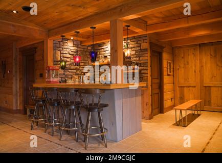 Voll ausgestattete graue Holzbar mit antiken braunen Holzbarhockern im Familienzimmer im Erdgeschoss im flachen Holzprofil und Holzhaus. Stockfoto