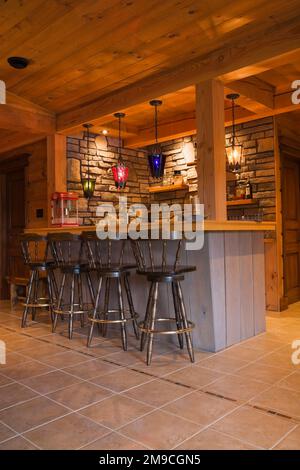 Voll ausgestattete graue Holzbar mit antiken braunen Holzbarhockern im Familienzimmer im Erdgeschoss mit flachem Holzprofil und Holzhaus. Stockfoto