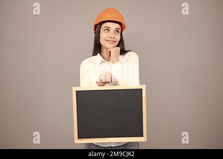 Junge lustige Frau Bauleiterin. Frau Baumeister isoliertes Porträt mit Schutzhelm und Brett. Grauer Hintergrund, leerer Speicherplatz, Kopierbereich. Stockfoto