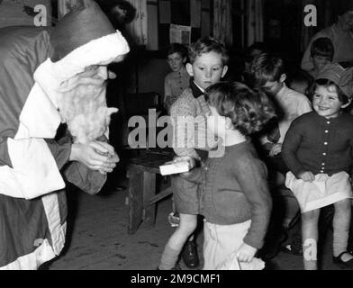 Mädchen und Jungen warten darauf, den Weihnachtsmann zu sehen, der Geschenke verteilt; er spricht intensiv mit einem kleinen Mädchen in einer Strickjacke Stockfoto