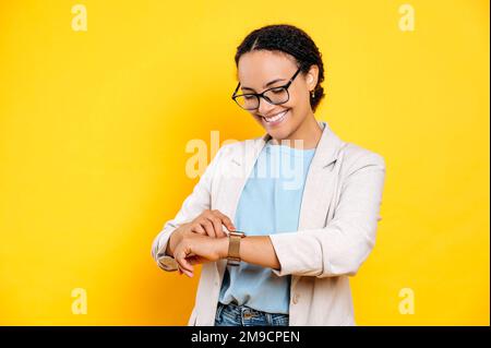 Positiv angenehm stilvoll gekleidete brasilianerin oder Latino-Frau, Geschäftsfrau, schaut auf die Uhr an seinen Händen, prüft die Zeit, lächelt, steht auf isoliertem gelbem Hintergrund Stockfoto