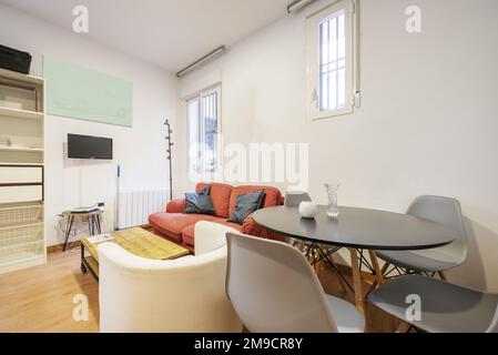 Loft-Apartment mit rundem schwarzem Esstisch und grauen Harzstühlen mit Holzfüßen und Zweisitzer-Sofa mit Couchtisch im Industriedesign Stockfoto