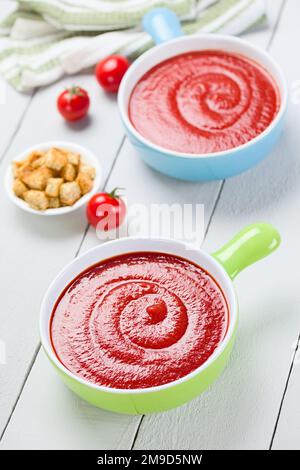 Frische hausgemachte Tomatensuppe in bunten Schüsseln, Ganzkorncroutons an der Seite, fotografiert auf weißem Holz (Selective Focus) Stockfoto