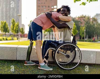 Zwei Männer mit Behinderungen umarmen sich in einem Stadtpark und repräsentieren Einheit und Integration trotz ethnischer Unterschiede und vielfältiger Fähigkeiten. Bild hoch Stockfoto