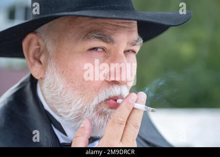 Nahaufnahme eines lustigen, stilvollen Großvaters im Ledermantel, der draußen raucht. Stockfoto