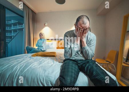 Streit um ein verheiratetes Paar. Der Mann sitzt auf dem Bett und hält seinen Kopf mit den Händen. Ehefrau oder wütende Freundin, die am anderen Ende des Bettes schräg sitzt. Beziehungsprobleme. Stockfoto