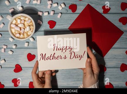 HAPPY MOTHER's DAY Text auf Karte Beschriftung positive Zitat Satz weibliche Hände halten valentinskarte roter Umschlag mit weißer Tasse Kaffee und Marshmallows auf blauem Holz Hintergrund. Romantische kleine Herzen Valentinstag. Feiertagmorgen. Ansicht von oben, flach liegend, minimalistisches Branding-Konzept Stockfoto