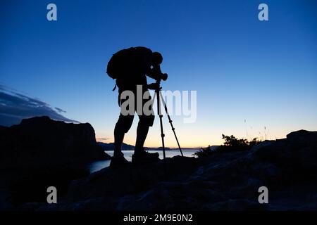 Silhouette des Fotografen auf dem Gipfel des Berges bei Sonnenuntergang. Naturfotograf in Aktion. Analoge Filmfotografie Stockfoto