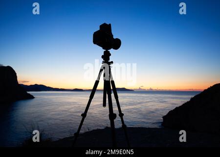 Fotokamera Silhouette auf Stativ am felsigen Strand mit wunderschönem Sonnenuntergang im blauen Meer auf Meereslandschaft Hintergrund. Analoge Filmfotografie Stockfoto
