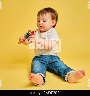 Ihr Kind spielt mit lustigen Rasselgeräuschen auf einem Stock auf dem gelben Hintergrund des Studios. Fröhlicher Kindermusiker, der Schlaginstrumente spielt. Kind A Stockfoto