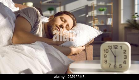 Mann, der auf dem Sofa am Tisch schläft, mit Wecker um 6 Uhr morgens Stockfoto