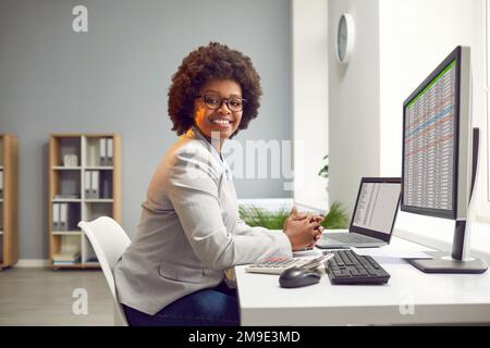 Glückliche lächelnde afroamerikanische Geschäftsfrau, Buchhalterin, die an einem Laptop mit Tischen im Büro arbeitet. Stockfoto