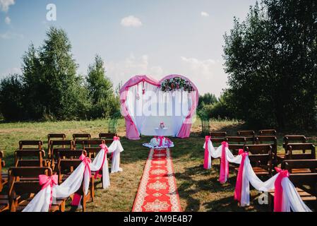 Hochzeitsbogen und Stühle für die Gäste für die Zeremonie, dekoriert mit weißen und pinkfarbenen Stoffen und Blumen Stockfoto