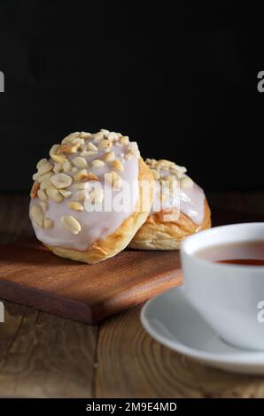 Leckere süße glasierte Brötchen mit Erdnüssen und einer Tasse Tee auf einem Holztisch Stockfoto