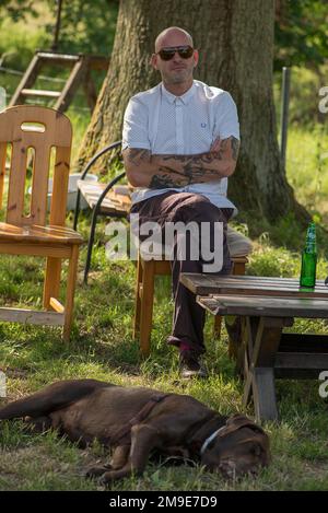 Junger Mann mit Tätowierungen und Sonnenbrille, ein schlafender Labrador davor, Mecklenburg-Vorpommern, Deutschland Stockfoto