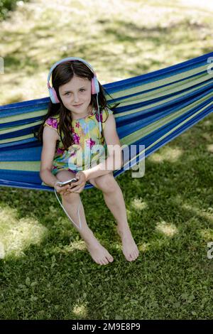 Mädchen, 8 Jahre, sitzt in einer Hängematte im Schatten und hört Musik mit Kopfhörern Stockfoto