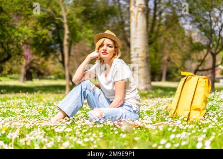 Ein junges blondes Mädchen mit Hut genießt den Frühling in einem Park in der Stadt, Urlaub in der Natur und neben Gänseblümchen Stockfoto