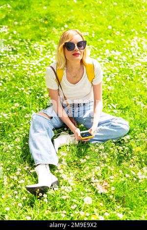 Porträt einer blonden Touristin mit Hut und Sonnenbrille, die im Frühling neben Gänseblümchen auf dem Gras sitzt, in die Kamera schaut und die Natur genießt Stockfoto