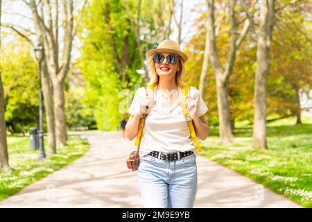 Eine blonde Touristin, die mit Hut und Sonnenbrille lächelt und im Frühling in einem Park in der Stadt mit der Kamera spaziert und die Sonne genießt Stockfoto