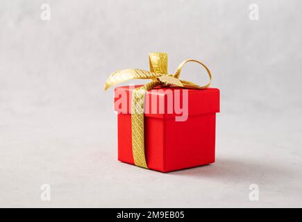 Eine rote Geschenkbox mit einem goldenen Band auf grauem Hintergrund. Das Konzept der Urlaubsfotografie. Überraschung zum Valentinstag, Geburtstag, Hochzeit. Kopieren Sie Spac Stockfoto
