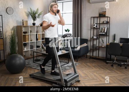 Portrait des gesunden Sportlers, der mit seinem Smartphone spricht, während er zu Hause Cardio-Training macht. Blonder, bärtiger Mann in weißem T-Shirt, der auf dem Laufband joggt. Stockfoto