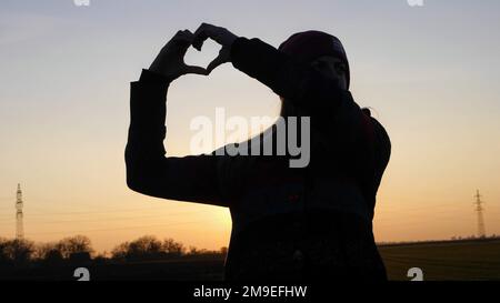 Die Silhouette einer Frau, die bei Sonnenuntergang mit den Händen am Himmel ein Herz formt Stockfoto