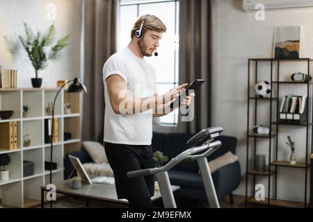 Seitliches Porträt eines jungen, aktiv bärtigen, kaukasischen Geschäftsmanns im Sport- und Headset-Training zu Hause, der mit einem Tablet arbeitet und abends auf dem Laufband Kardio-Übungen macht. Stockfoto