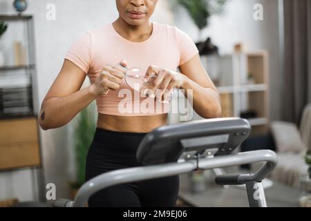 Verkürztes Porträt der Hände mit einer Flasche Wasser einer aktiven afroamerikanischen sportlichen Frau in Sportbekleidung Training zu Hause, Laufen, Herz-Kreislauf-Training, auf Laufband. Stockfoto